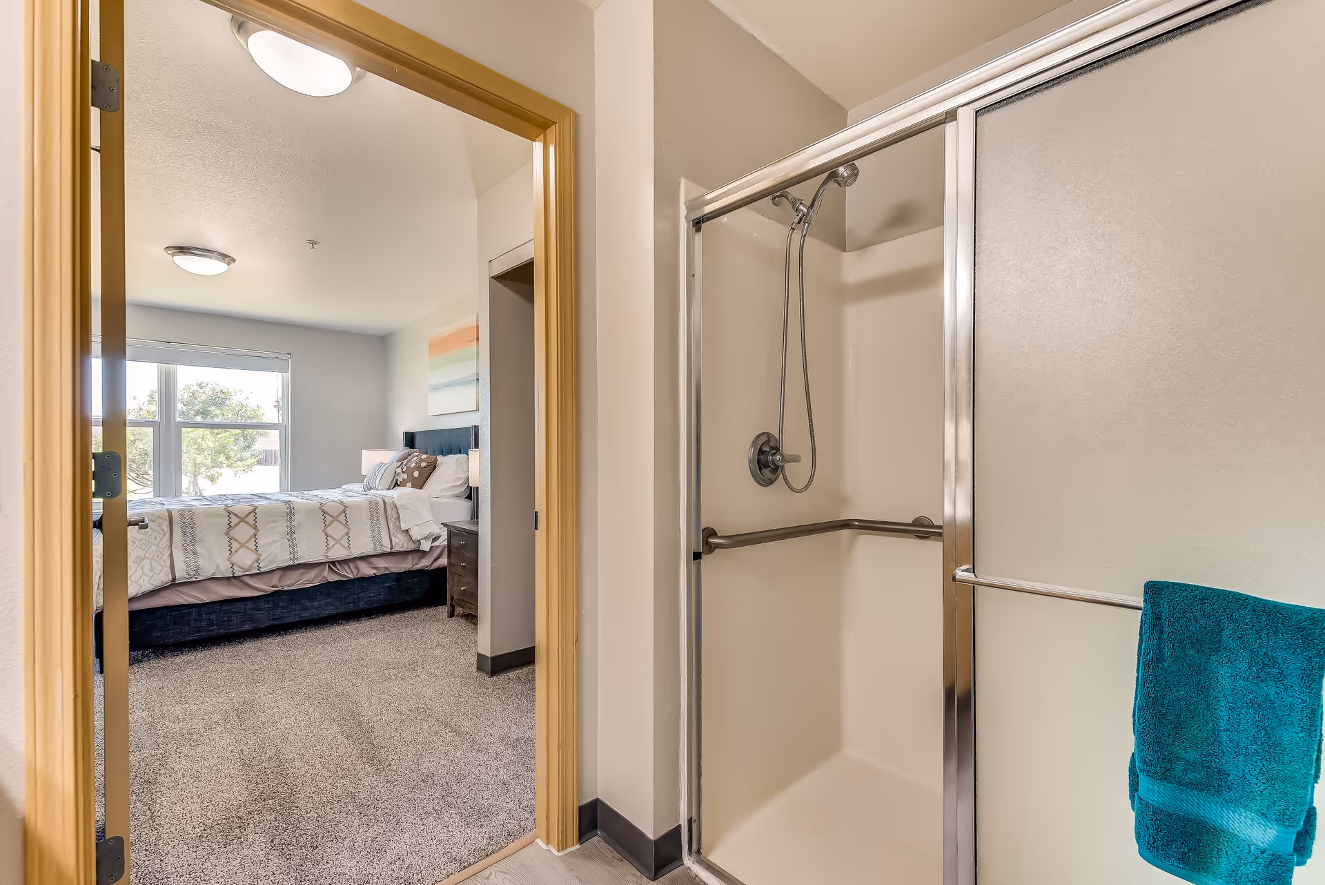 View of a bathroom with a shower stall featuring a sliding glass door and a handheld showerhead. A teal towel hangs on a bar outside the shower. Through an open doorway, a bedroom is visible with a bed, nightstand, and a large window letting in natural light.