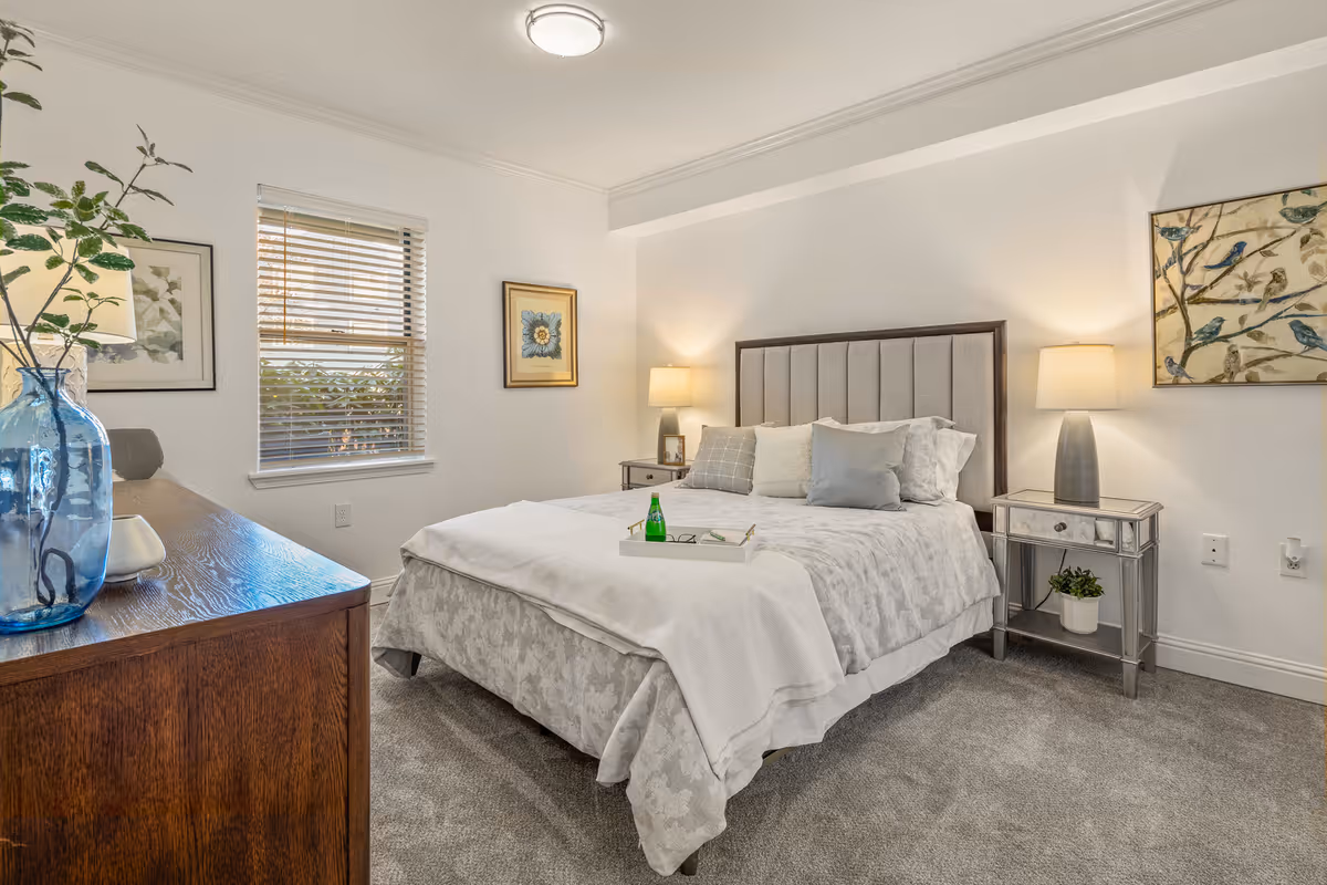A bright, neatly staged bedroom with a bed featuring an upholstered headboard, nightstands with lamps, a dresser, window and wall art.