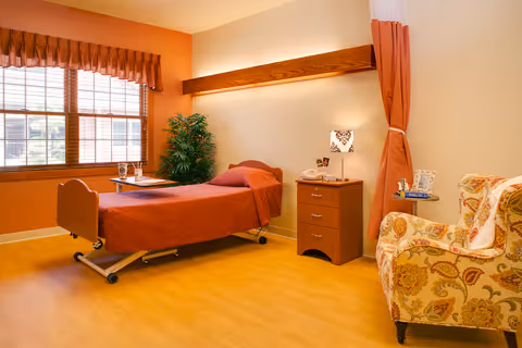 A well-lit room with a single hospital-style bed covered in a rust-colored blanket, a wooden nightstand with a lamp and some items on it, a patterned armchair, a green potted plant, and a window with wooden blinds and a rust-colored valance.