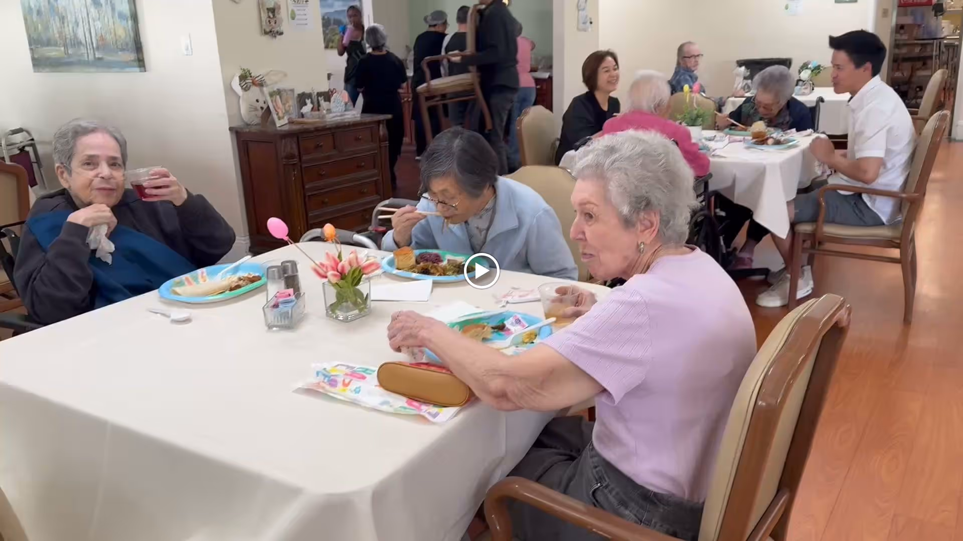 A group of elderly people sitting around tables in a dining room, eating and drinking. There are plates of food, napkins, and a small flower vase on the table. Some people are engaged in conversation while others are focused on their meals. The room has wooden flooring and light-colored walls with some decorations.