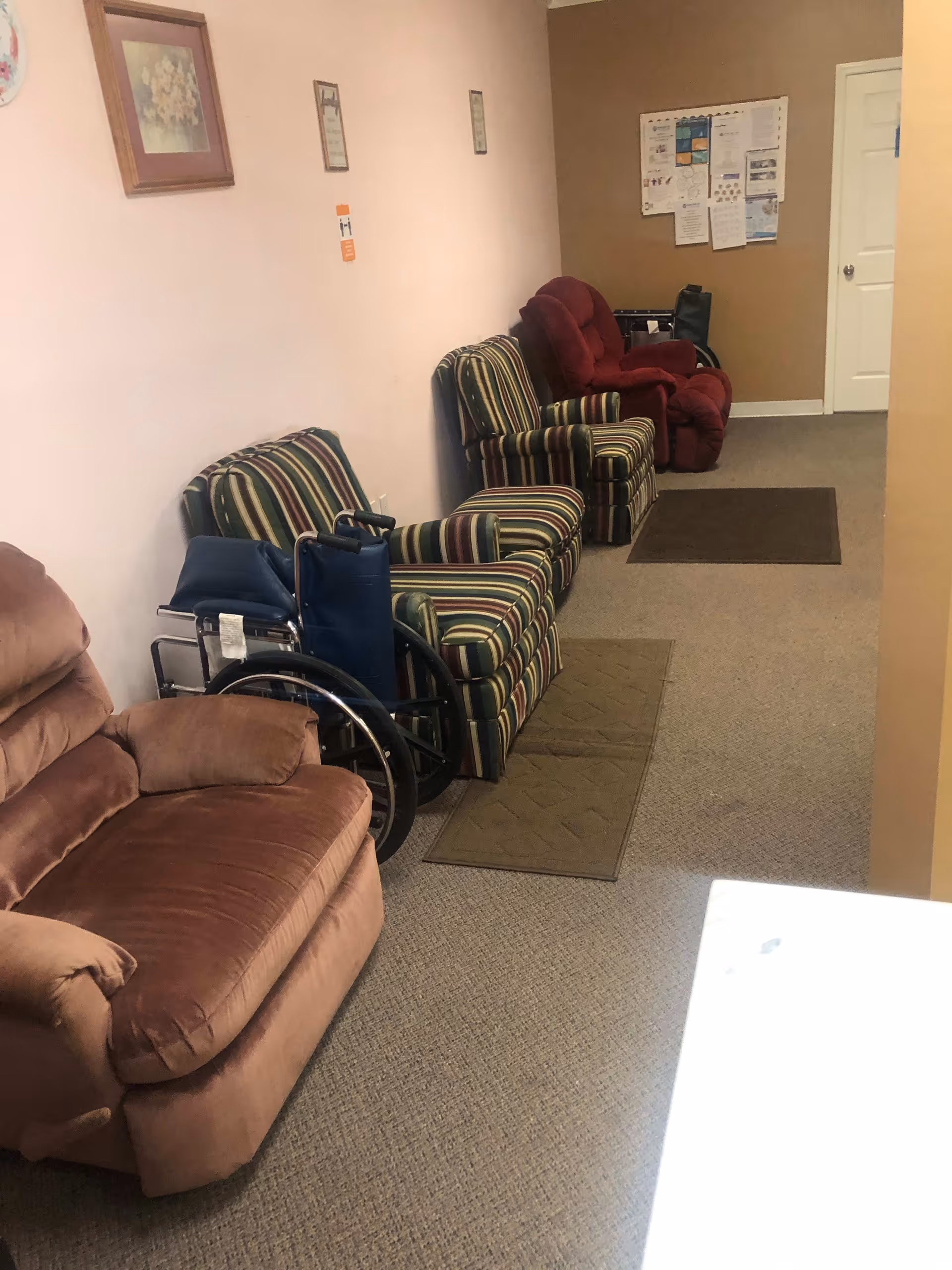 A hallway with several upholstered chairs lined up against a wall, including a brown recliner, two striped armchairs, and a red recliner. A wheelchair is positioned between the brown recliner and the striped chairs. The walls are decorated with framed pictures and notices, and there are small rugs on the carpeted floor.