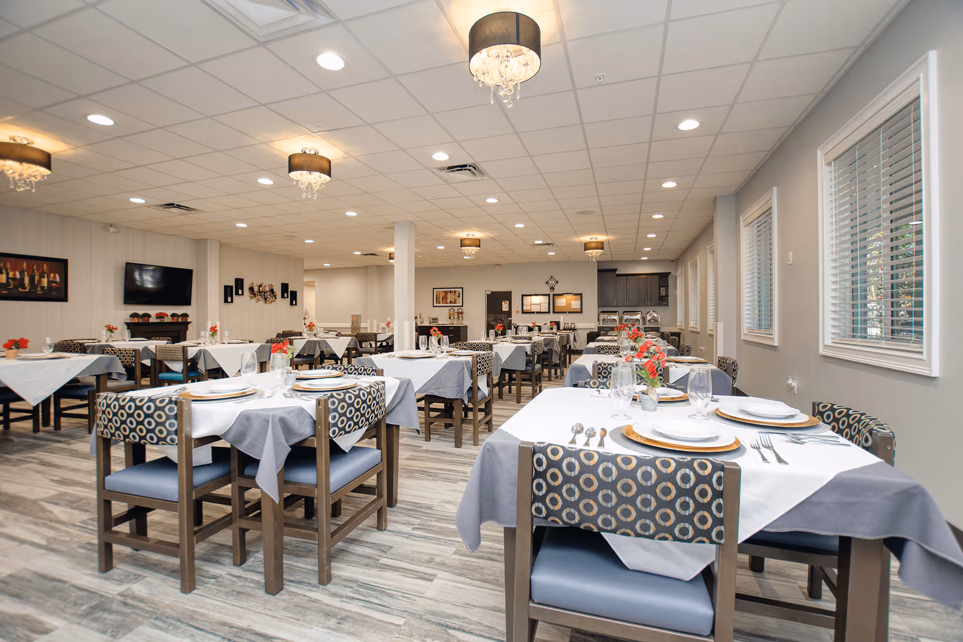 A spacious and well-lit dining room in a senior living facility with multiple tables covered with white and gray tablecloths, each set with plates, glasses, and silverware. The chairs have patterned upholstery and blue cushions. The room features large windows with blinds, modern ceiling lights with chandeliers, and a television mounted on the wall.