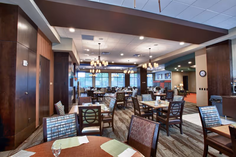 A spacious dining room in a senior living facility with multiple tables and chairs arranged neatly. The room features large windows allowing natural light, modern chandeliers hanging from the ceiling, and a patterned carpet. The walls have dark wood paneling and there is a clock on one wall.