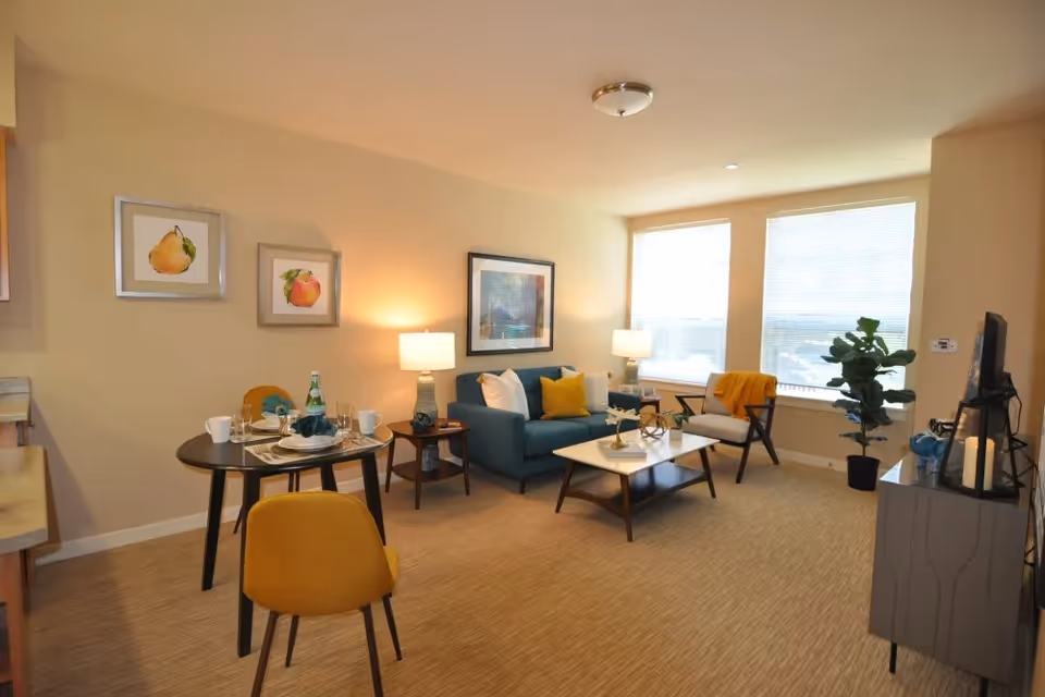 A cozy living room and dining area with beige walls and carpet. The living room features a blue sofa with white and yellow pillows, a white coffee table, a side table with a lamp, a modern armchair with a yellow throw, and a large window letting in natural light. The dining area has a small round table set for two with plates, glasses, and a bottle, accompanied by yellow chairs. There are framed fruit artworks on the wall and a TV on a stand with decorative items.