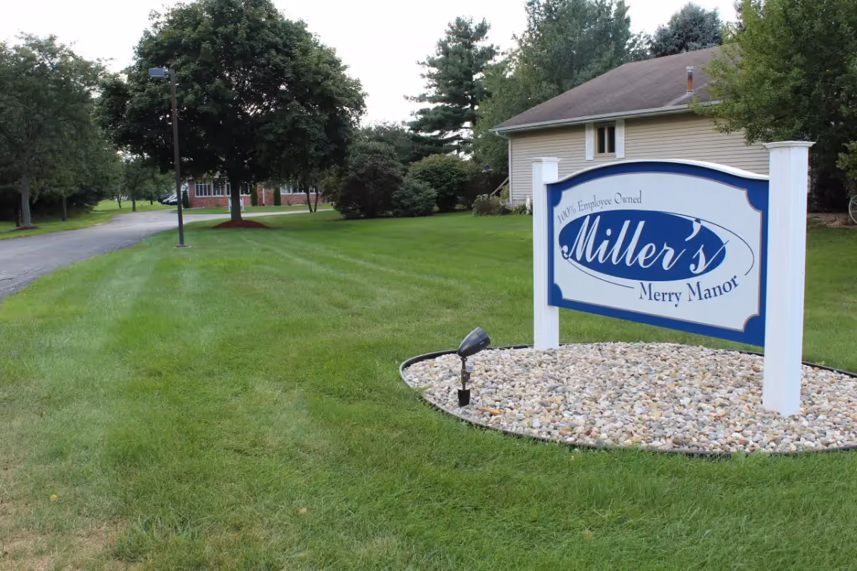 White and blue sign reading "Miller's Merry Manor" on a manicured lawn in front of a single-story building.