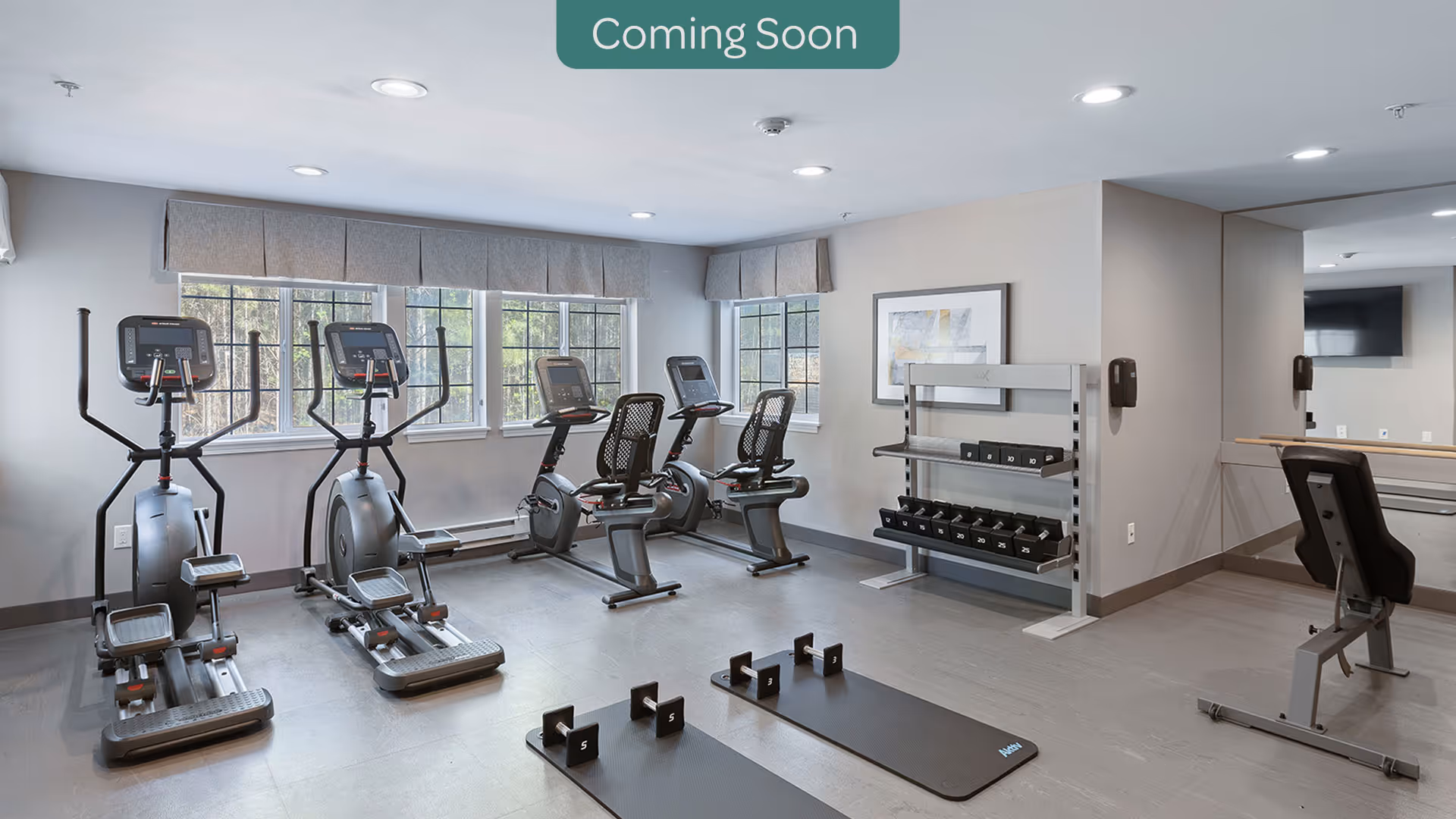 A modern fitness room with exercise equipment including two elliptical machines, two stationary bikes, a weight bench, dumbbells on a rack, and two exercise mats on the floor. The room has large windows with gray valances, light gray walls, and recessed ceiling lights.
