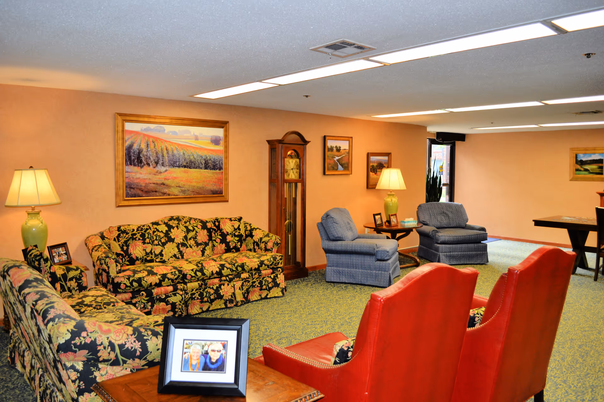 A cozy living room area with floral patterned sofas, two blue armchairs, and two red leather chairs. The walls are decorated with landscape paintings and a tall wooden grandfather clock. Two green table lamps with beige shades sit on side tables, and a framed photo of two elderly women is on a wooden table in the foreground. The room has a carpeted floor and fluorescent ceiling lights.