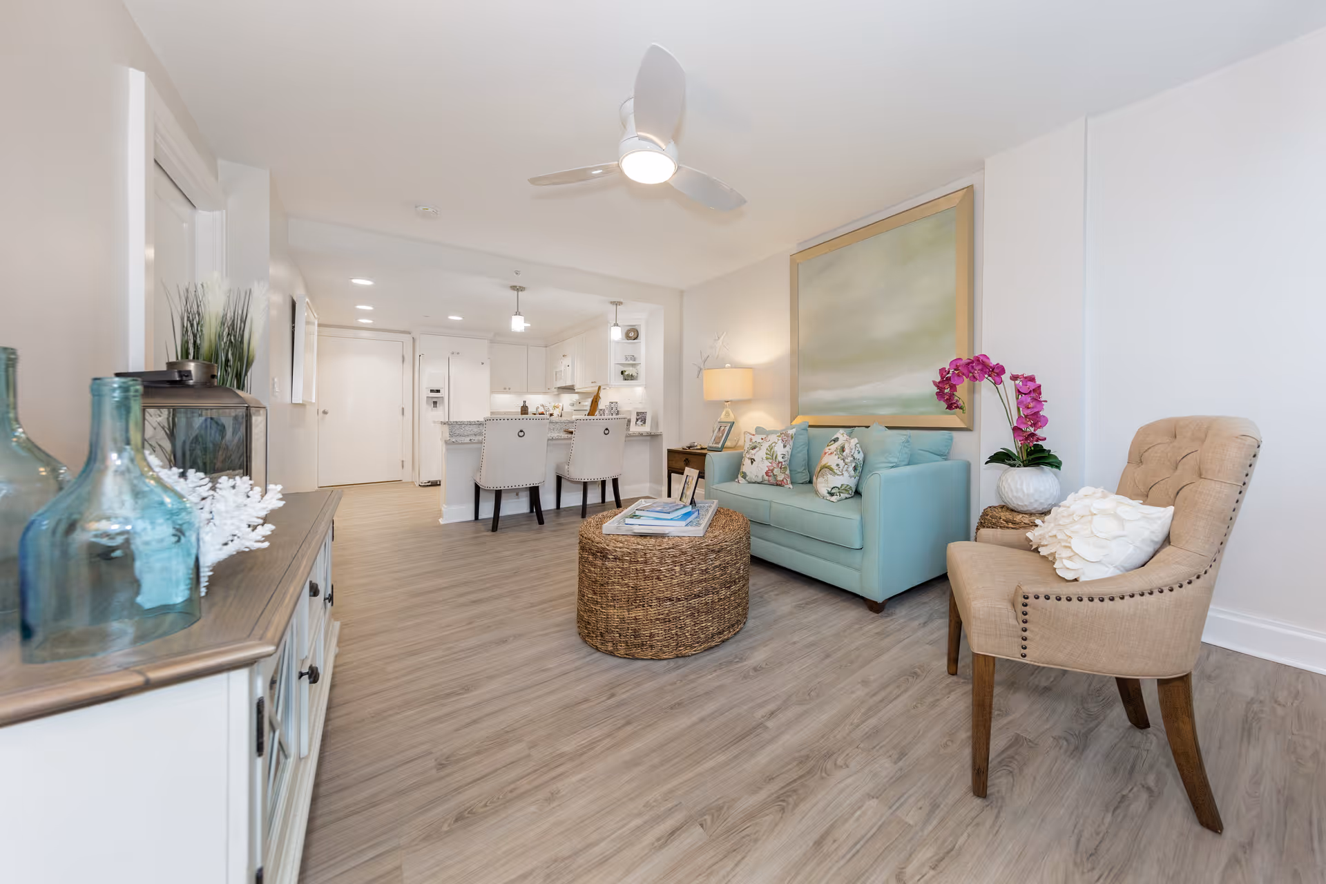 A bright and modern living room area with light wood flooring, a light blue sofa with floral pillows, a beige armchair with a white decorative pillow, a round woven coffee table with books on it, and a large abstract painting on the wall. The room opens into a white kitchen with a breakfast bar and two upholstered bar stools. Decorative glass bottles and plants are on a sideboard to the left.