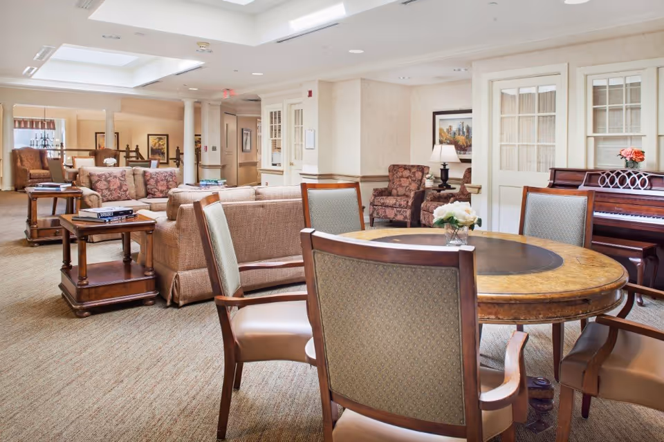 Spacious senior living lounge with a round table and chairs in the foreground, sofas and armchairs, side tables and a piano in the background.