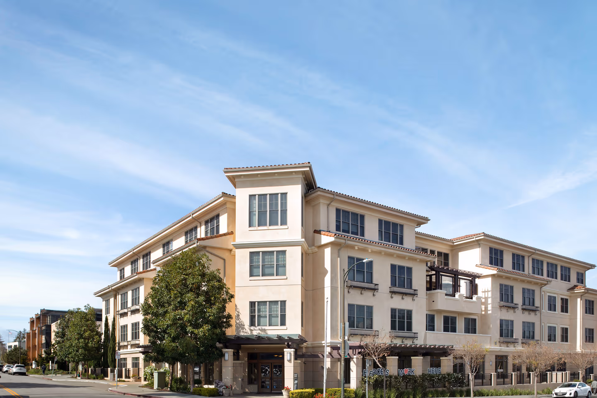 Exterior view of a multi-story senior living facility building with beige walls, multiple windows, and a tiled roof under a clear blue sky. Trees and shrubs are planted around the building, and a street with parked cars runs alongside it.