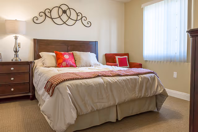 A neatly made bed with decorative pillows, a nightstand and lamp, wall decor, and a small chair by a window in a softly lit bedroom.