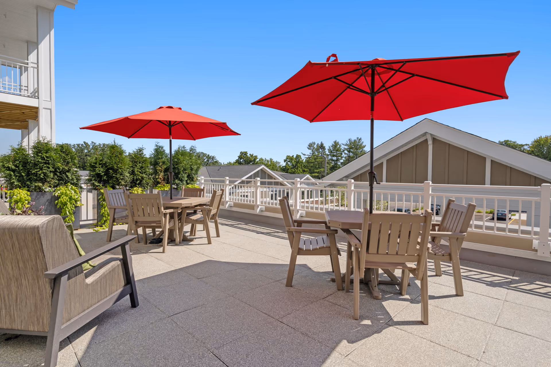 Rooftop patio with tables, wooden chairs and red umbrellas overlooking nearby buildings on a sunny day.