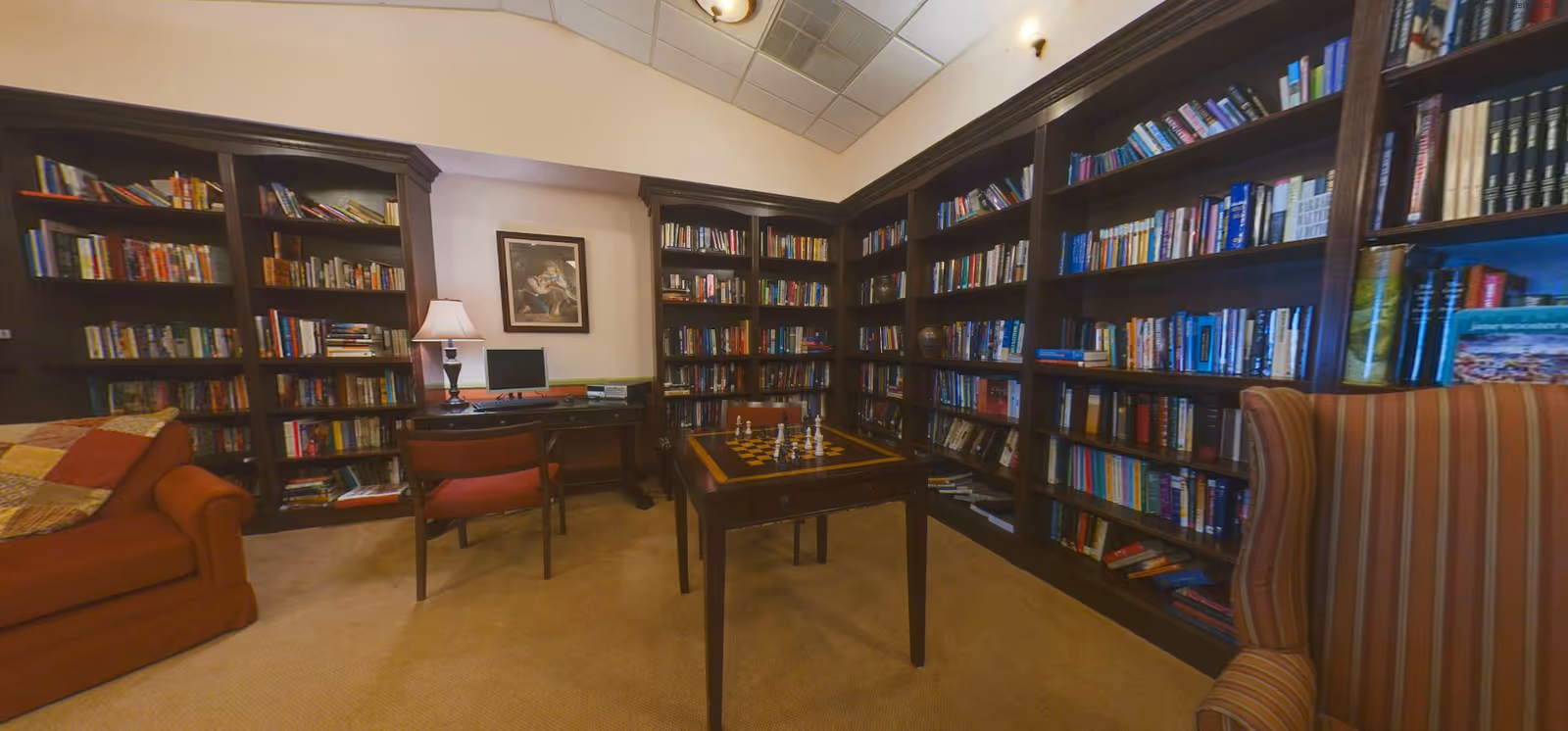 A cozy library room with dark wooden bookshelves filled with books lining the walls. There is a small table with a chessboard and chess pieces set up in the center of the room. A desk with a computer and a lamp is against the wall, accompanied by a chair. A striped armchair and a red sofa with a patchwork quilt are also visible. The room has a beige carpet and a high ceiling with recessed lighting.