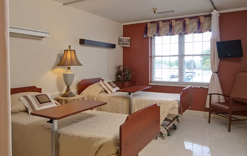 A well-lit room with two hospital-style beds, each with beige bedding and pillows, equipped with overbed tables. A lamp sits on a small table between the beds. There is a window with blinds and a floral valance letting in natural light. A brown armchair and a wall-mounted television are visible near the window. The walls are painted beige and a reddish-brown accent wall.