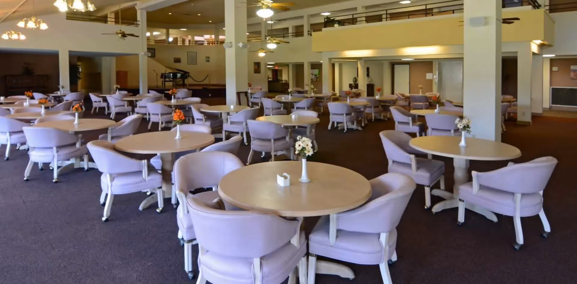 A spacious dining room with multiple round tables, each surrounded by four lavender cushioned chairs. Small floral centerpieces are placed on each table. The room features high ceilings with ceiling fans and overhead lighting. There is a piano in the background and a mezzanine level with railings overlooking the dining area.