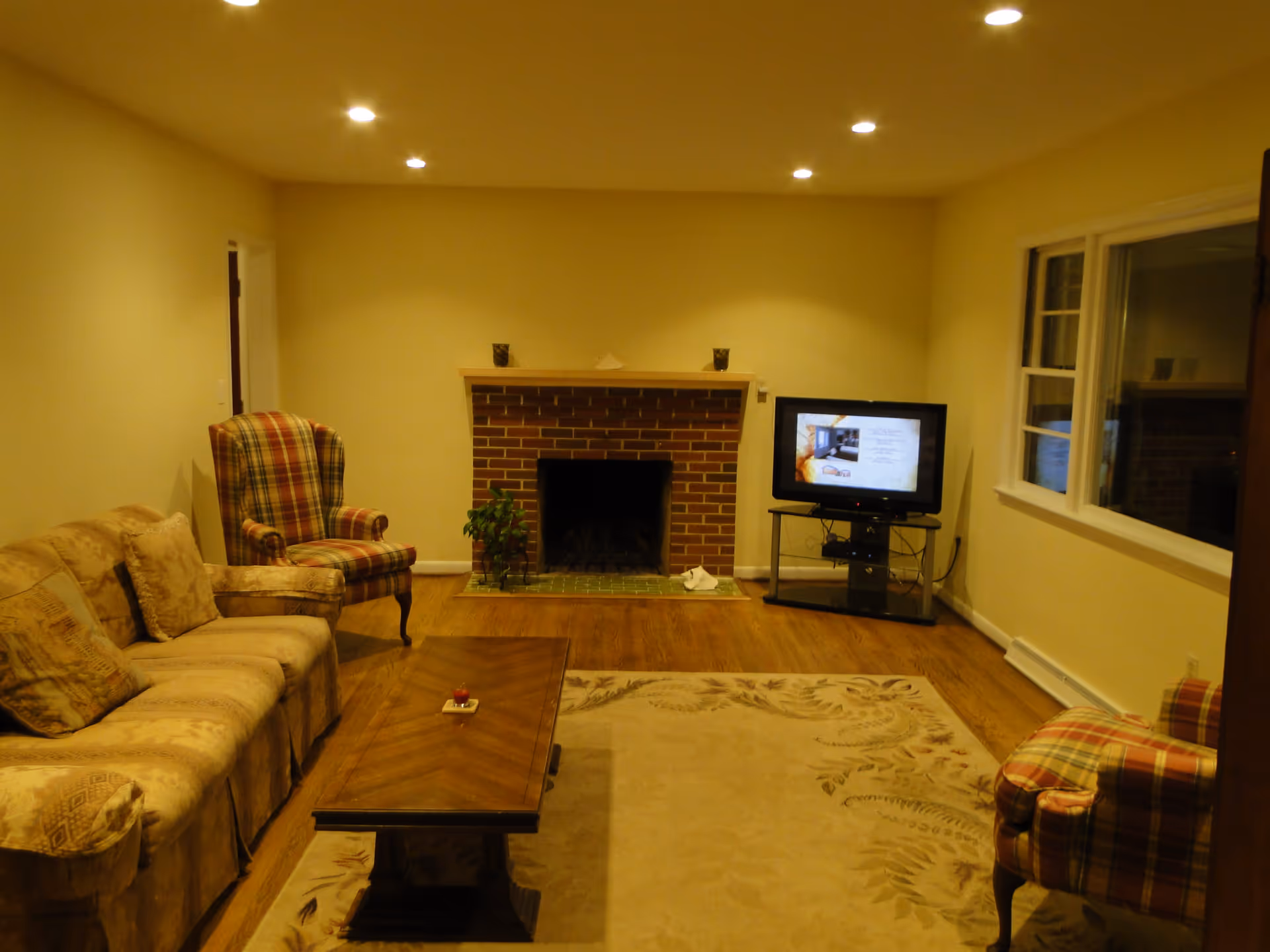 A cozy living room with a beige couch, two plaid armchairs, a wooden coffee table, a brick fireplace, a television on a stand, and a large window. The room has wooden flooring and recessed ceiling lights.