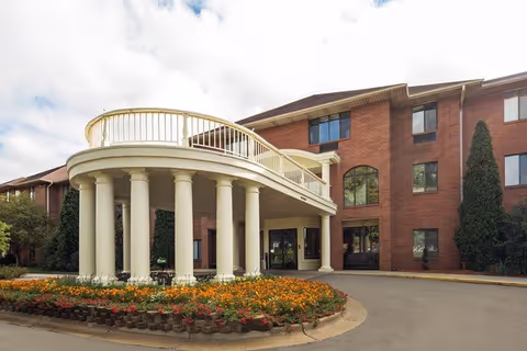 Exterior view of a senior living facility building with a circular driveway and a covered entrance supported by large white columns. The building is made of red brick with multiple windows and surrounded by landscaped flower beds and greenery.