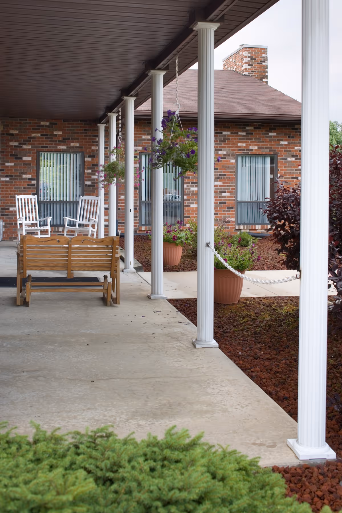 Covered front porch with white columns, hanging flower baskets, wooden benches and rocking chairs outside a brick building.