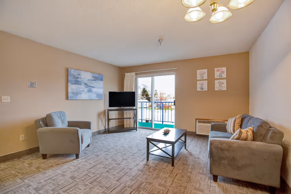 A bright living room with a sofa and armchair facing a coffee table and TV, plus a sliding glass door leading to a balcony.