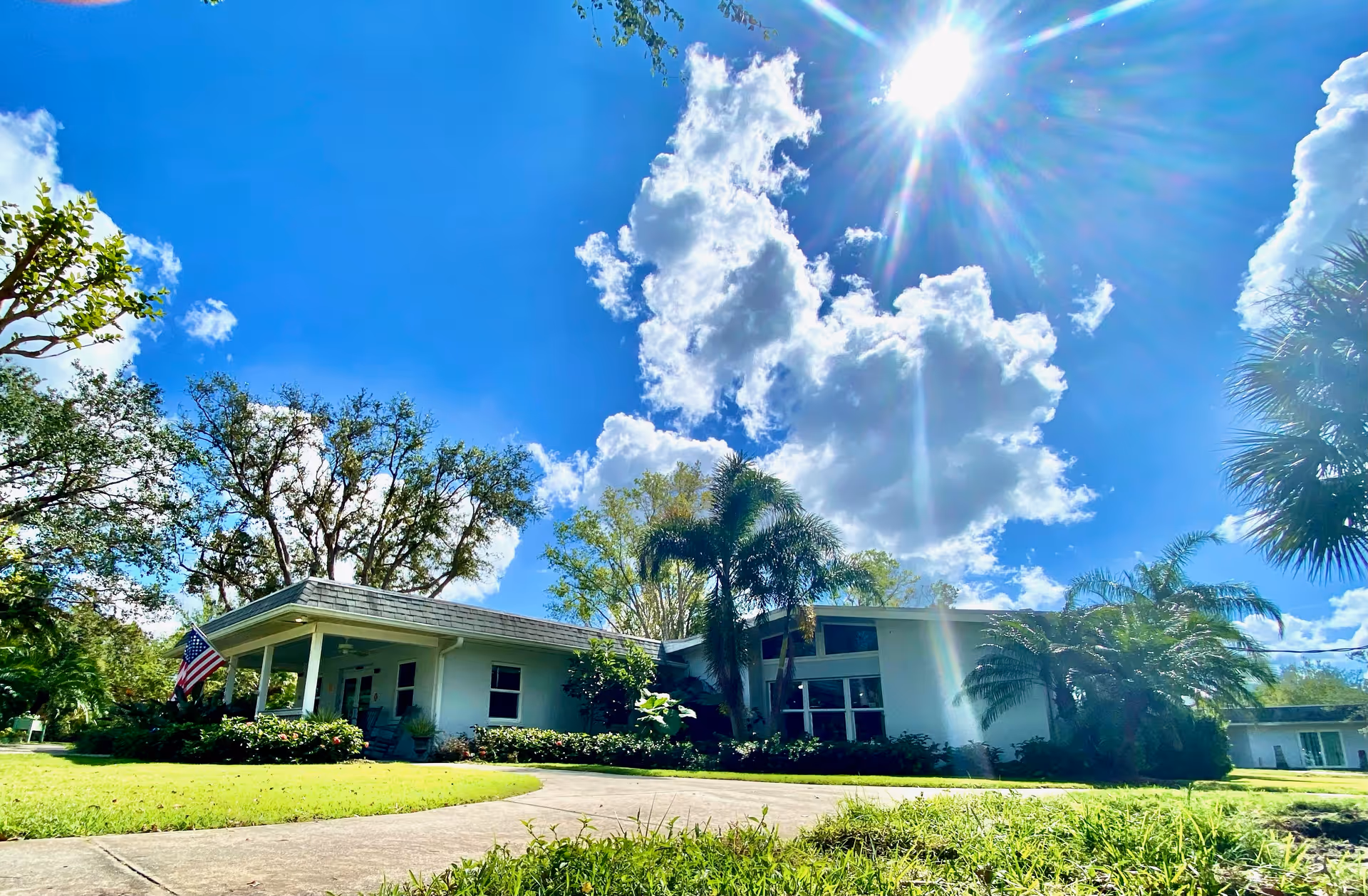 Front exterior of a single-story white senior living building with palm trees, a manicured lawn, an American flag, and a bright sunny sky.