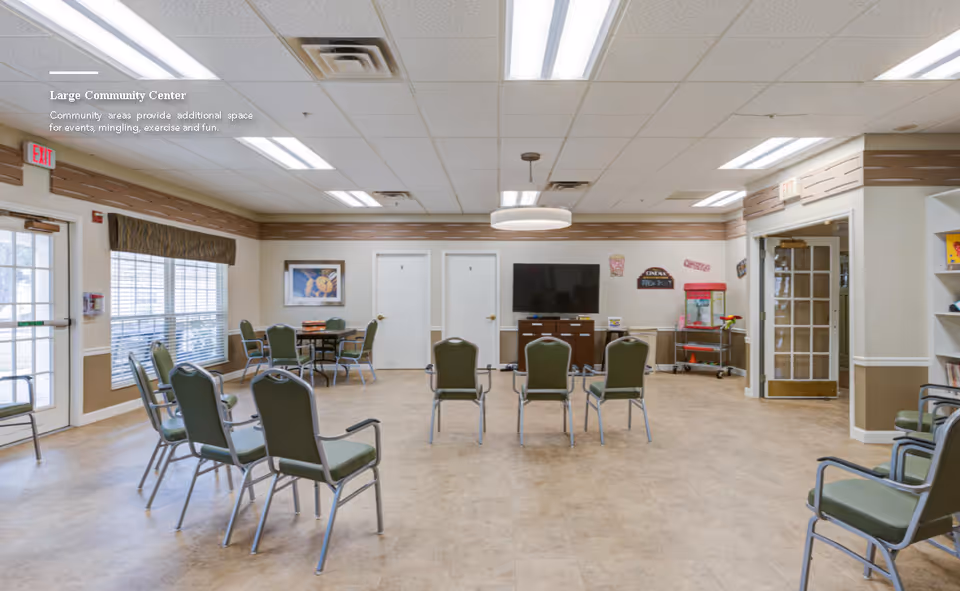 A large community center room with several green chairs arranged in rows facing a flat-screen TV mounted on a wooden cabinet. The room has beige walls with a brown trim, a tiled floor, and fluorescent ceiling lights. There are windows with blinds on the left side, a popcorn machine and movie-themed decorations on the right, and a round table with chairs near the windows.