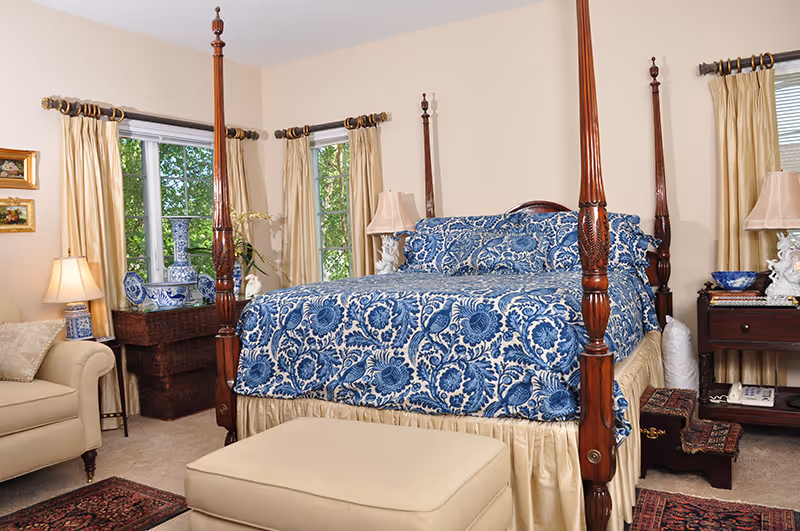 A bedroom with a large four-poster bed covered in blue and white floral bedding. The room has beige walls and carpet, two windows with gold curtains, a beige armchair, a beige ottoman, two bedside tables with lamps, decorative blue and white vases, and a small wooden step stool next to the bed.