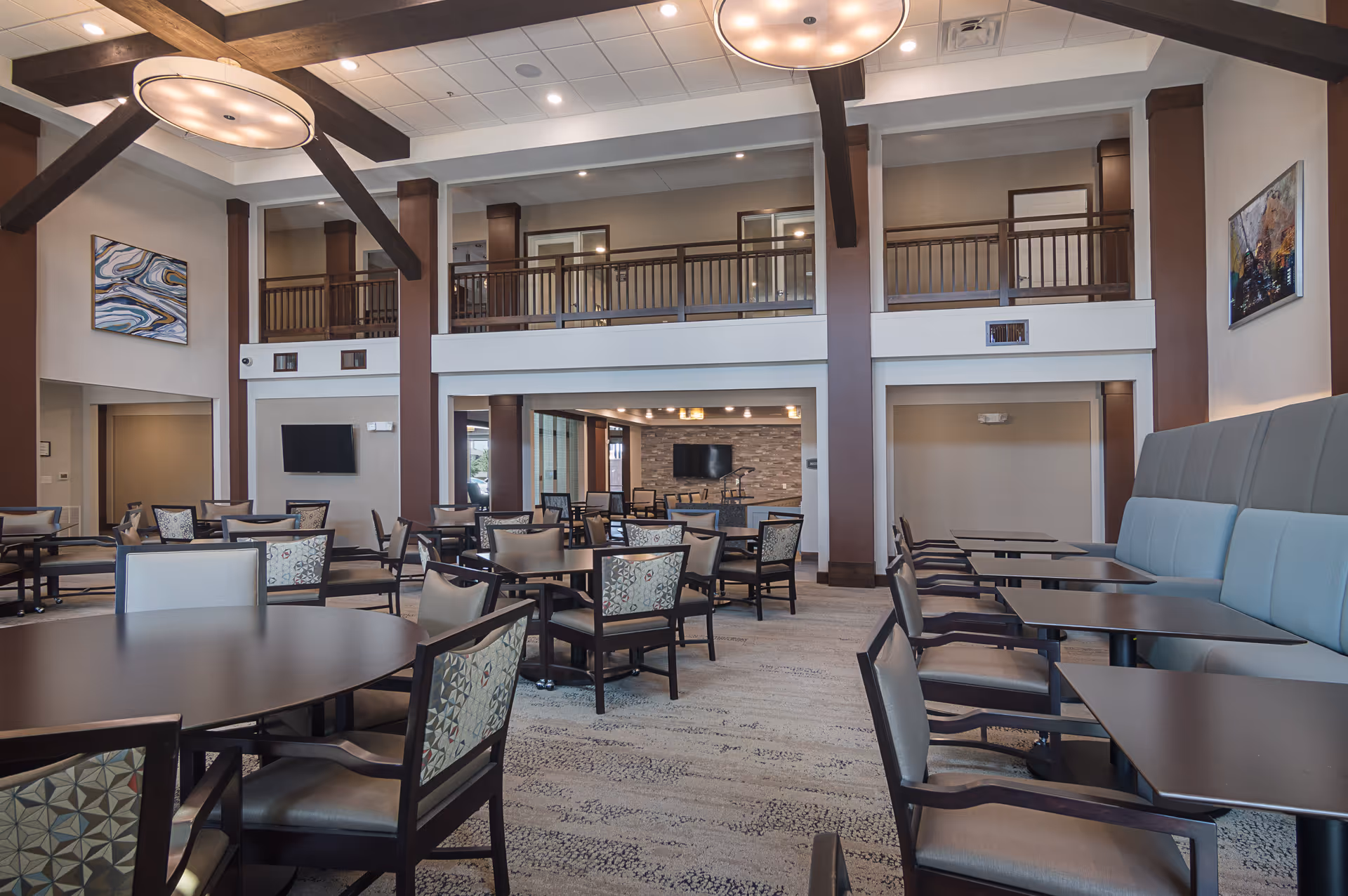 Spacious dining area with multiple tables and chairs arranged neatly. The room features high ceilings with exposed wooden beams and modern circular light fixtures. There is a balcony railing on the upper level and two flat-screen TVs mounted on the walls. The walls are decorated with abstract paintings, and the floor is carpeted with a patterned design.