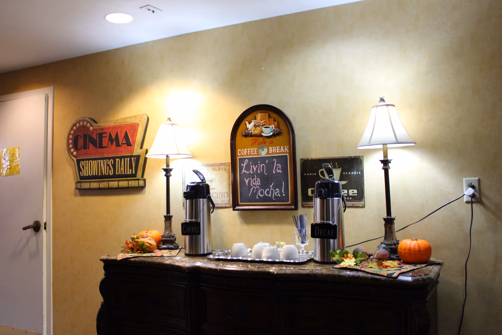 Indoor coffee station with two lamps, coffee urns, seasonal pumpkins and vintage wall signs on a sideboard.