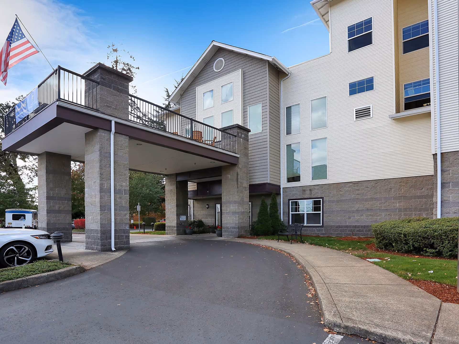Covered entrance/porte-cochère and front façade of a senior living building with an American flag and a parked car.