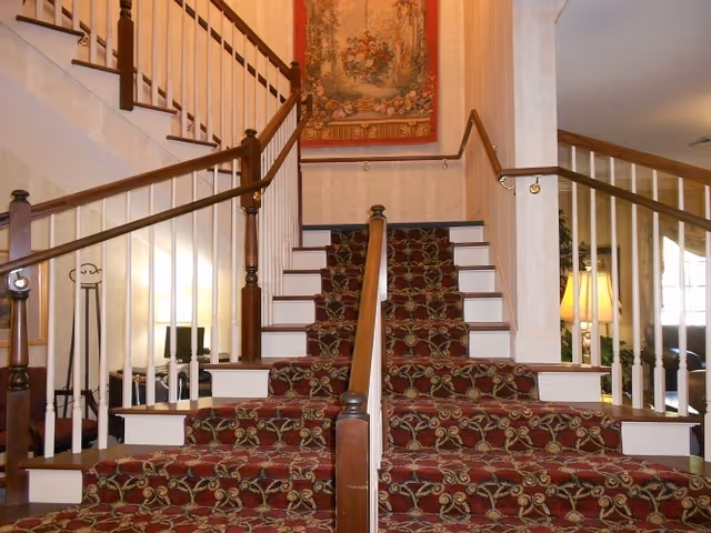 A carpeted staircase with a floral pattern splits into two directions at a landing. The staircase has wooden handrails and white balusters. A tapestry with a floral design hangs on the wall above the landing. To the right, a lamp and some furniture are partially visible in a living area.