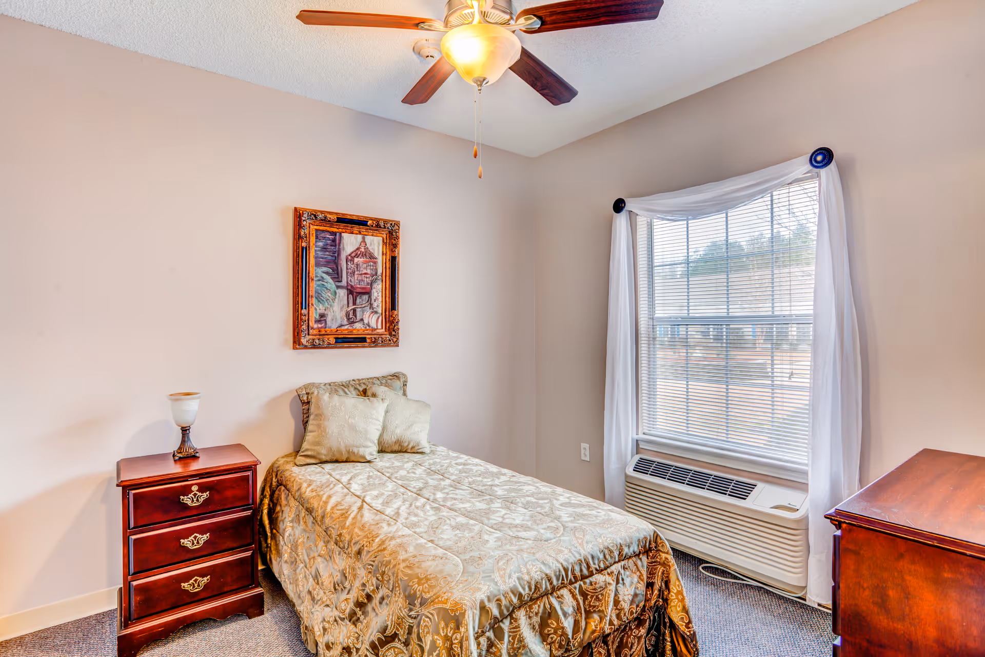 A small bedroom with a single bed covered in a gold patterned bedspread and two matching pillows. Next to the bed is a wooden nightstand with three drawers and a lamp on top. Above the bed hangs a framed painting. A window with white curtains and blinds is on the right wall, with an air conditioning unit installed below it. A ceiling fan with a light fixture is mounted on the ceiling.