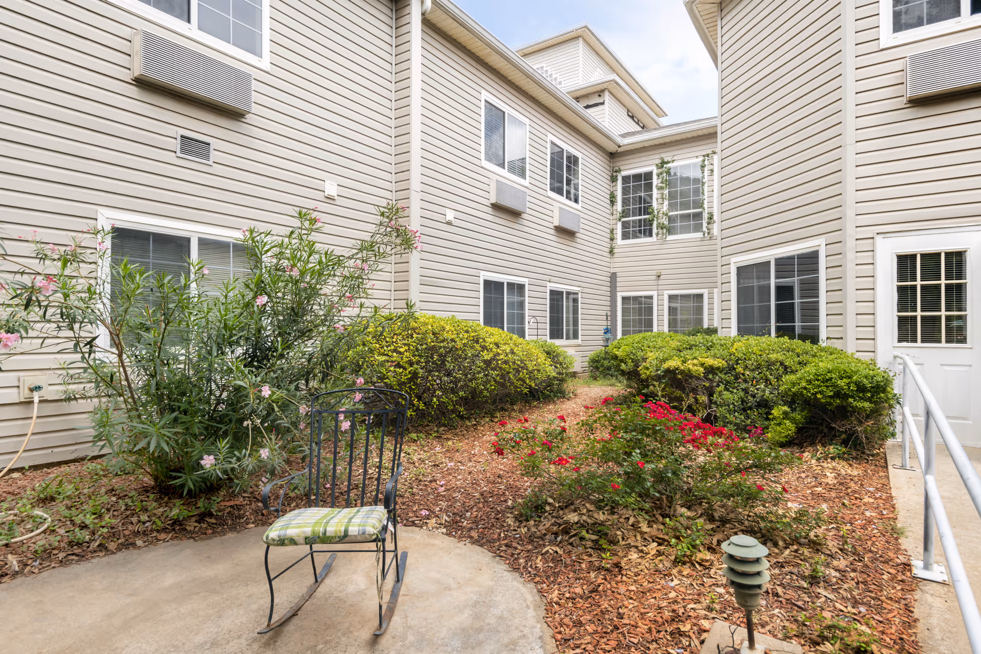 Landscaped courtyard at a senior living facility with a metal chair, shrubs, and surrounding beige siding building.