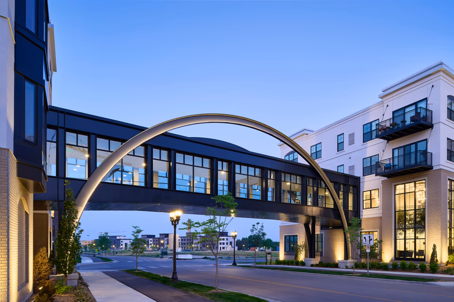 Exterior view of a modern senior living facility at dusk featuring a large illuminated archway connecting two buildings with multiple windows and balconies. The street below is lined with small trees and street lamps.
