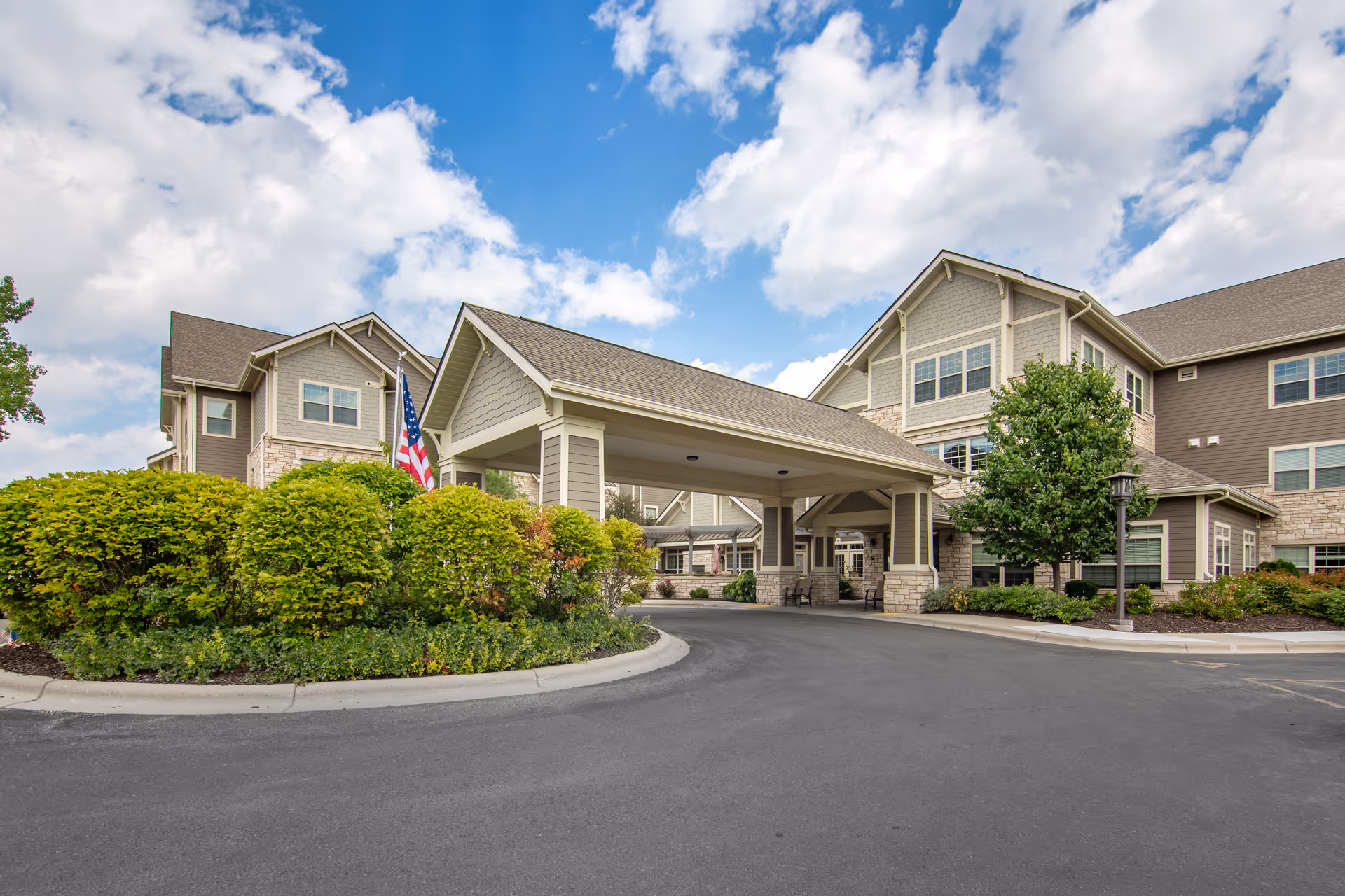 Exterior view of Brookdale Madison West senior living facility showing a covered entrance with stone and siding facade, surrounded by greenery and an American flag under a partly cloudy blue sky.