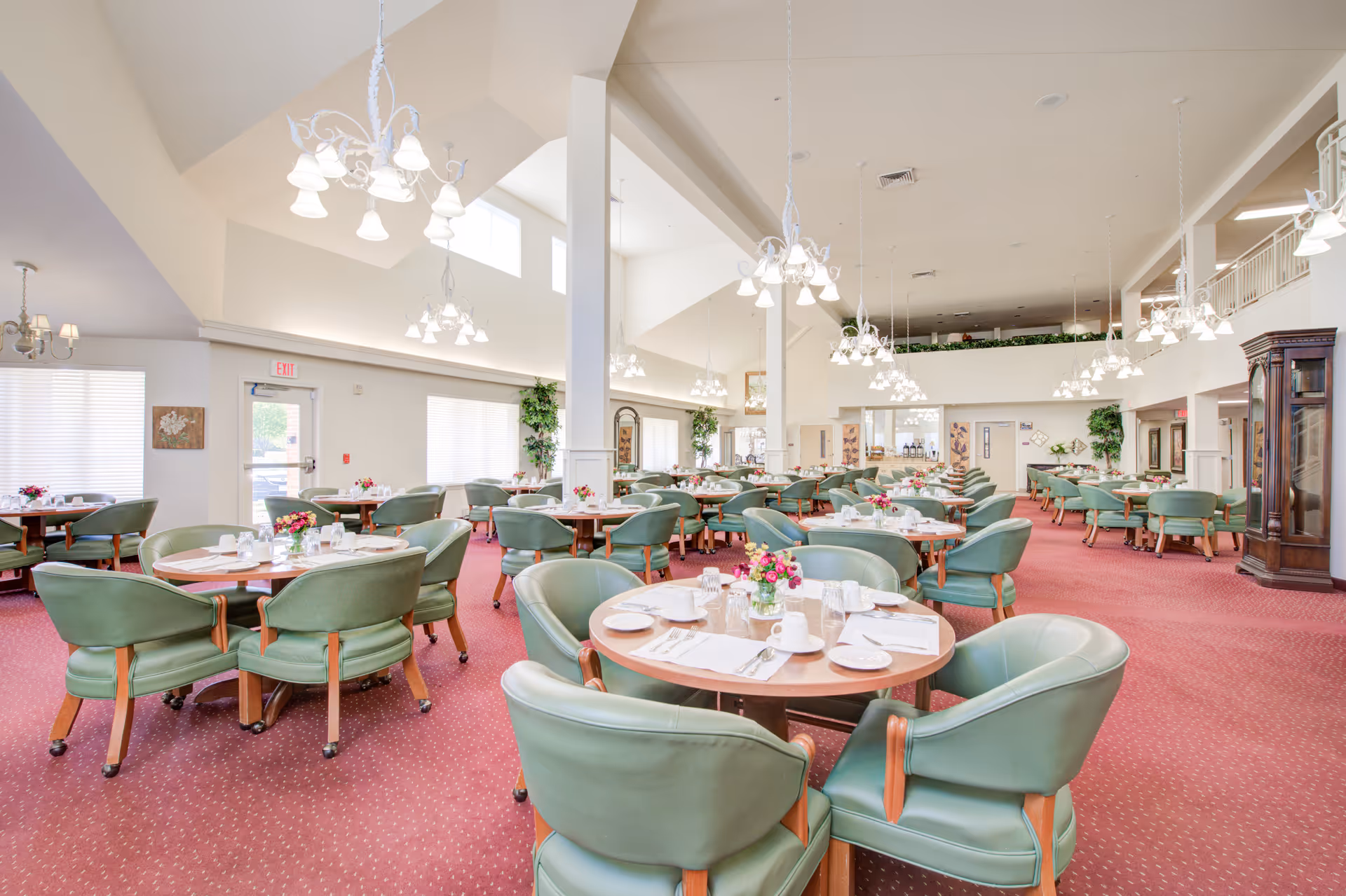 A spacious senior living dining room with round wooden tables set with white plates, cups, and silverware. Each table has green cushioned chairs and a small floral centerpiece. The room features high ceilings with multiple chandeliers, large windows letting in natural light, red carpet flooring, and decorative plants along the walls.