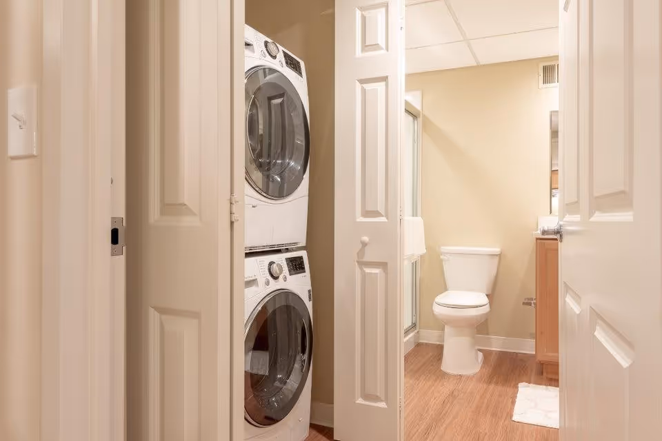 View through an open door into a bathroom with a toilet, a wooden vanity with a sink, and a shower with a glass door. Adjacent to the bathroom is a closet with stacked washer and dryer units. The walls are beige and the floor is wood.