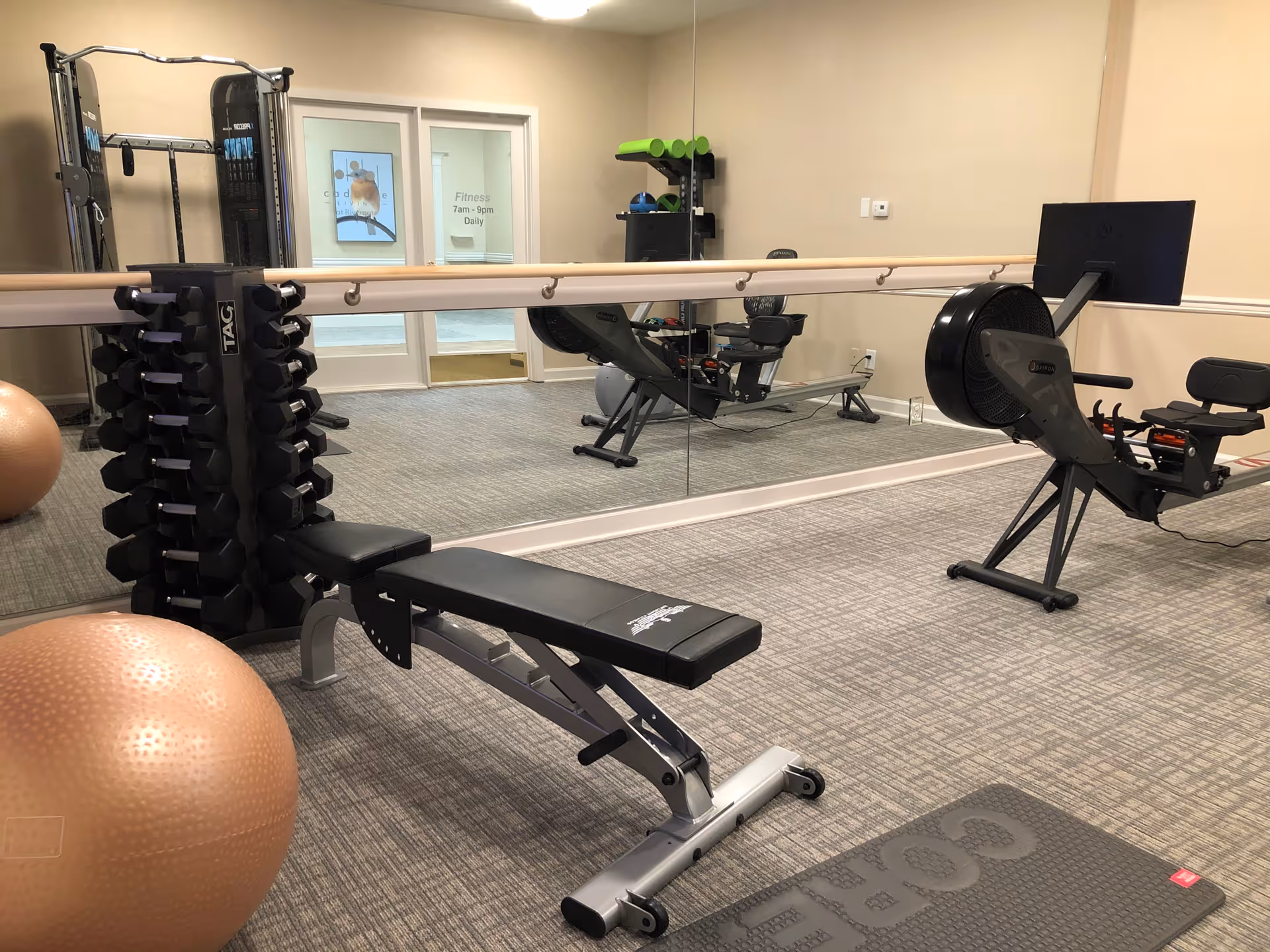 A fitness room with exercise equipment including a rowing machine, a weight bench, a rack of dumbbells, a large exercise ball, and a yoga mat. The room has a mirrored wall and a ballet barre along the mirror. There is a door with a sign indicating fitness hours from 7am to 9pm daily.