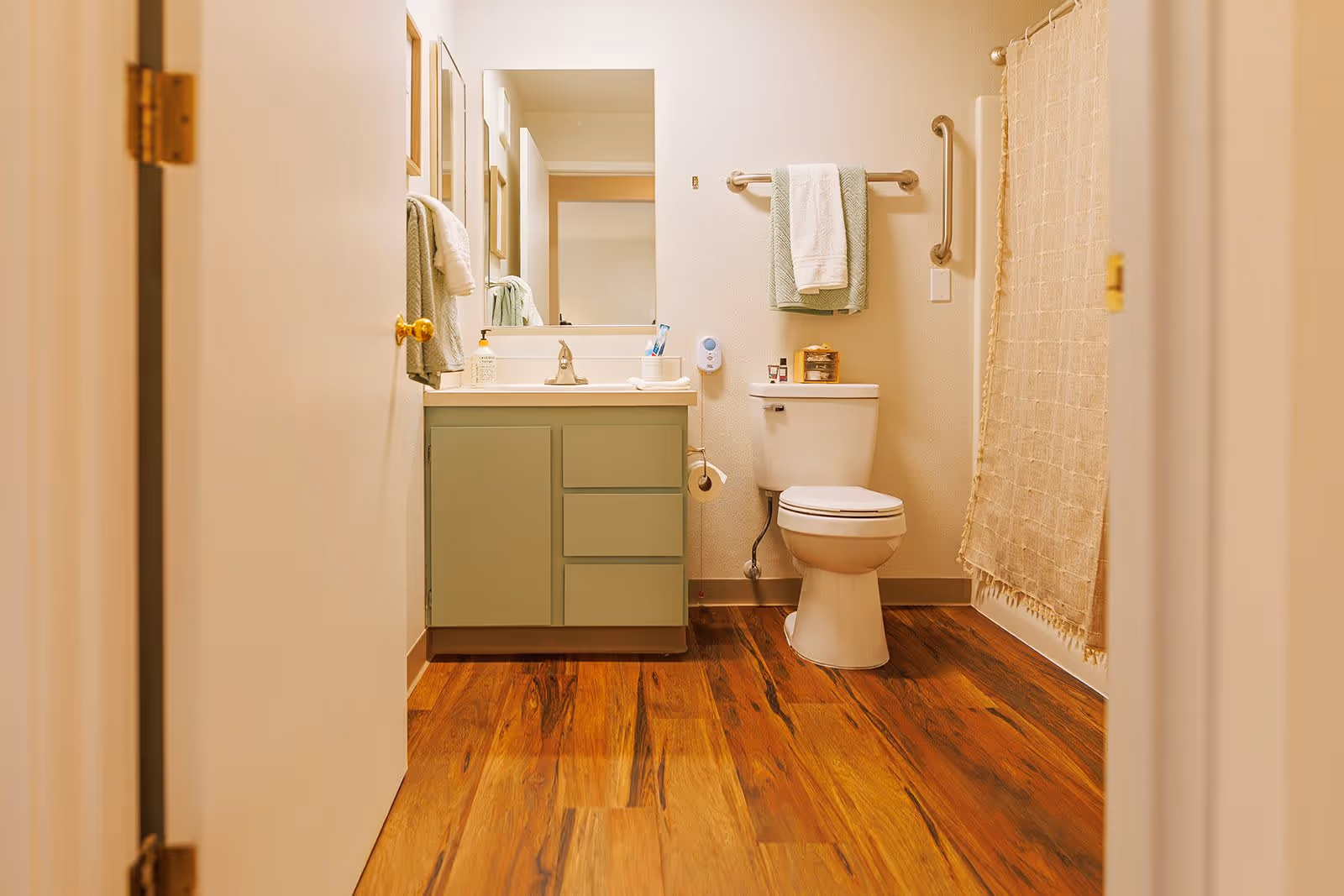 View into a bathroom with wooden flooring, a green vanity with a sink and mirror, a toilet with a towel rack above it holding two towels, and a shower with a beige curtain. The bathroom has light-colored walls and a partially open door.