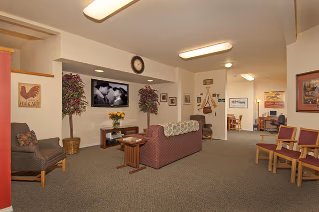 A cozy living room area in a senior living facility with a maroon couch, armchairs, and wooden chairs arranged around a TV mounted on the wall showing a black and white movie scene. The room has beige walls, carpeted floor, framed pictures, two potted plants, a clock above the TV, and a small desk with a computer in the background.