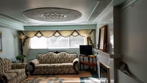 A living room with a patterned sofa and armchair, a small wooden table holding a flat-screen TV, a fireplace with a mirror above it, and a large window with decorative curtains allowing natural light to fill the room. The ceiling features an ornate circular design.