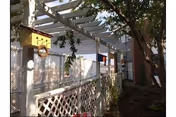 Outdoor area of a facility with a white lattice fence, a pergola overhead, a yellow birdhouse attached to a tree, and some greenery including trees and bushes.