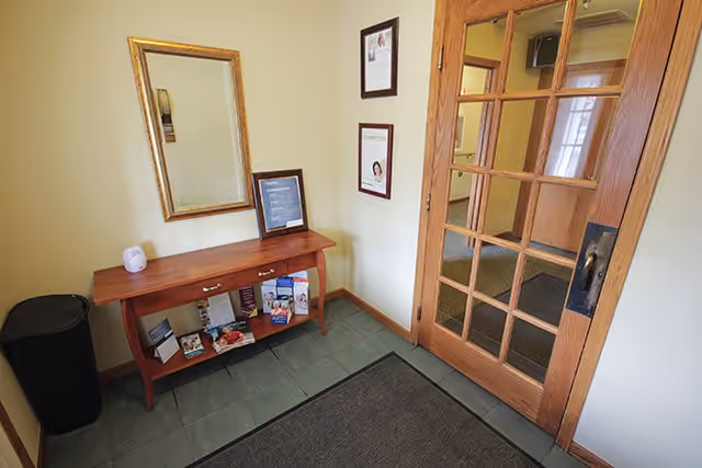 Small entryway area with a wooden console table holding brochures and a framed sign, a gold-framed mirror above it, a black trash bin to the left, and a wooden door with glass panes on the right leading to another room.