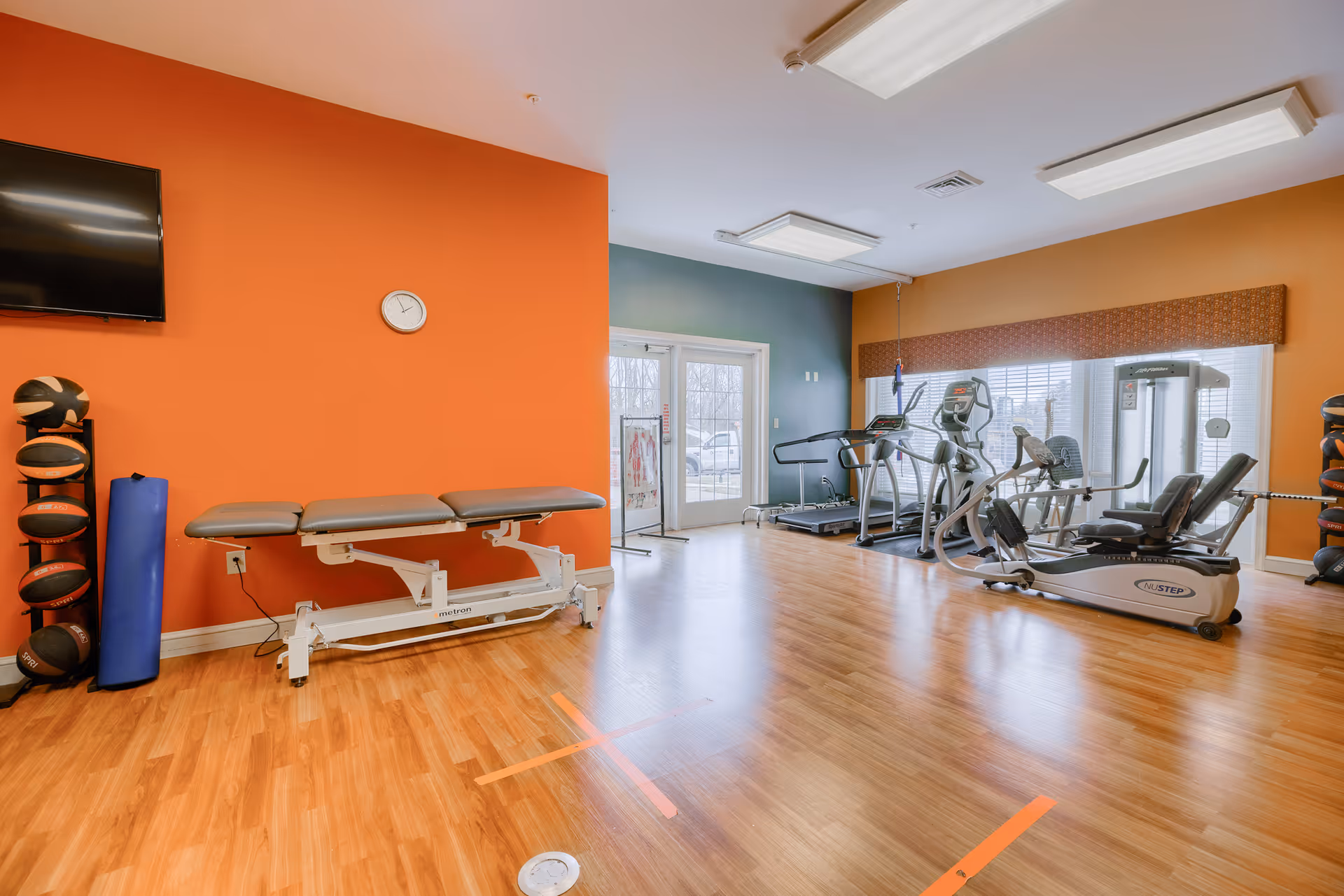 A fitness room with wooden flooring and orange and teal walls. The room contains exercise equipment including a treadmill, stationary bike, and weight machine. There is a padded exercise bench against the orange wall, a rack of medicine balls, a blue foam roller, a wall clock, and a mounted TV. Large windows and a glass door allow natural light into the room.