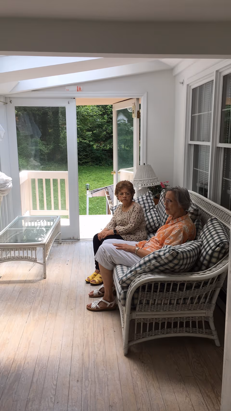 Two elderly women sitting on a white wicker couch with plaid cushions in a sunroom with wooden floors. The sunroom has large windows and a glass door that opens to a green outdoor area. There is a glass-top wicker coffee table and a lamp on a side table next to the couch.
