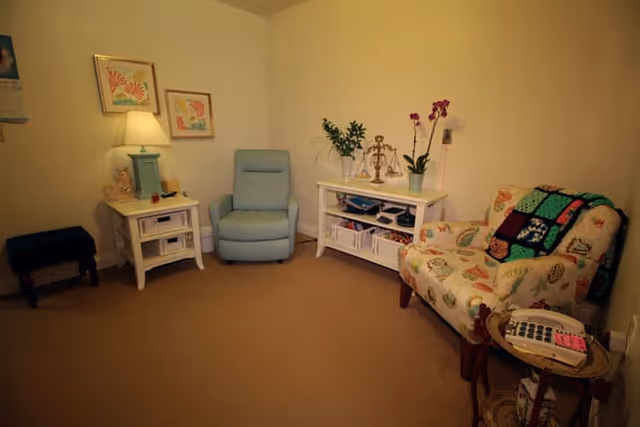 A cozy living room with a light blue armchair, a floral patterned armchair with a colorful quilt draped over it, a white side table with a lamp and decorative items, a white console table with plants and decor, and a small round table with a telephone. The walls are light-colored and there are framed artworks hanging on the wall.