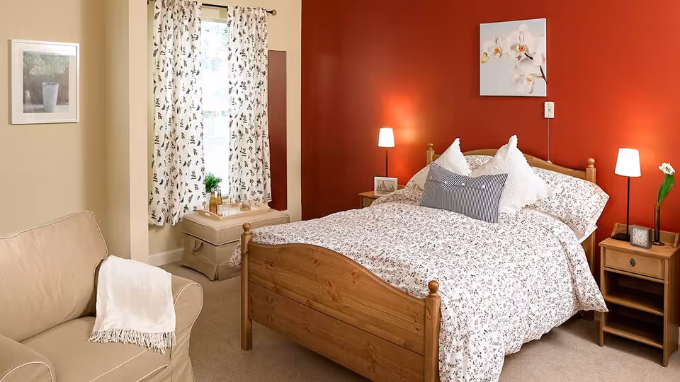 A cozy bedroom with a wooden bed frame and floral bedding. Two bedside tables each have a lamp, and one has a small clock and a flower vase. A beige armchair with a white throw blanket is positioned near a window with patterned curtains. A framed picture hangs on the wall beside the window, and a floral painting is above the bed on a red accent wall.
