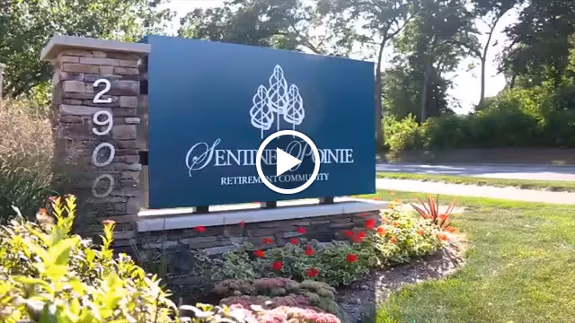 Outdoor view of a large sign for Sentinel Pointe Retirement Community, mounted on a stone base with the address number 2900 on the left side, surrounded by landscaping with flowers and greenery, with trees and a road in the background.