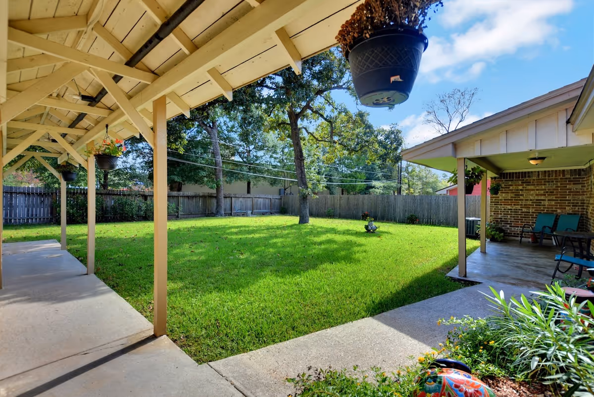 A sunny backyard with a green lawn, trees, and a wooden fence. There is a covered patio area with hanging flower pots and outdoor seating. The sky is blue with some clouds.