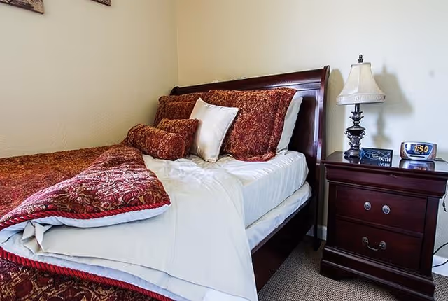 A neatly made bed with a dark wooden headboard, adorned with red and white pillows and a red patterned comforter. Next to the bed is a dark wooden nightstand with a decorative lamp, a small clock displaying 7:50, and a small sign that says 'Faith'. The room has light-colored walls and carpeted flooring.