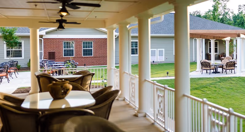 Covered outdoor patio area with tables and chairs, white columns supporting the roof, and a green lawn with another seating area under a pergola in the background.